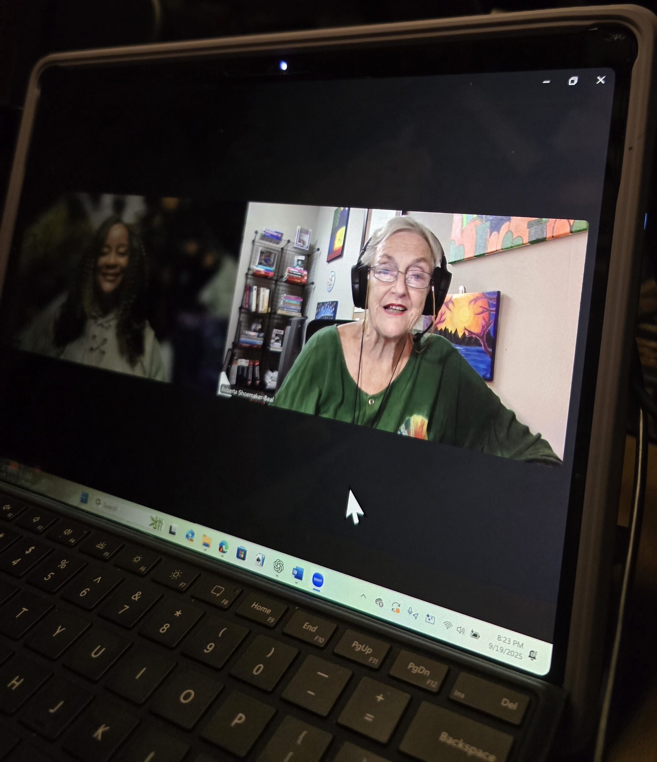A virtual meeting on a tablet screen shows two participants: a smiling woman with curly hair and an elderly woman in headphones, discussing topics with a colorful background.