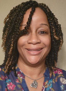 Smiling woman with shoulder-length twisted hair and a heart-shaped pendant, wearing a floral blouse, exudes warmth and confidence.