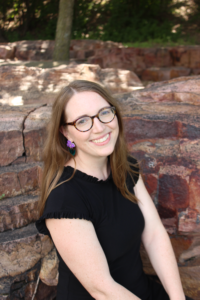 Woman with long hair and glasses smiles while sitting on a stone wall in a natural setting. Her purple earrings add a pop of color to her black dress.